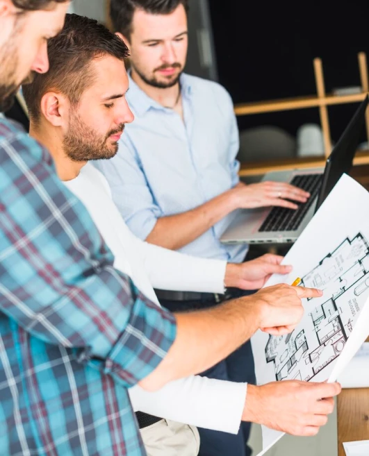 Three men reviewing blueprints in an office; one points at the plans, another listens, and a third works on a laptop.