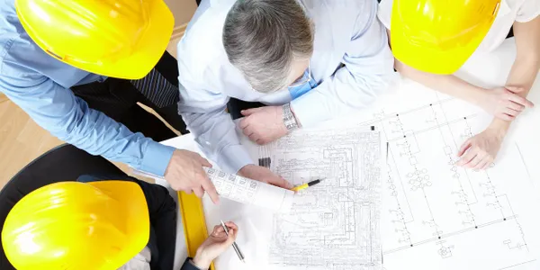 Engineers in yellow hard hats reviewing blueprints on a table.