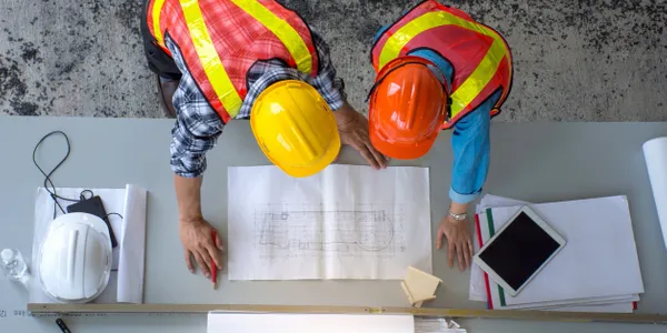 Overhead view of two engineers in safety vests reviewing blueprints on a table.