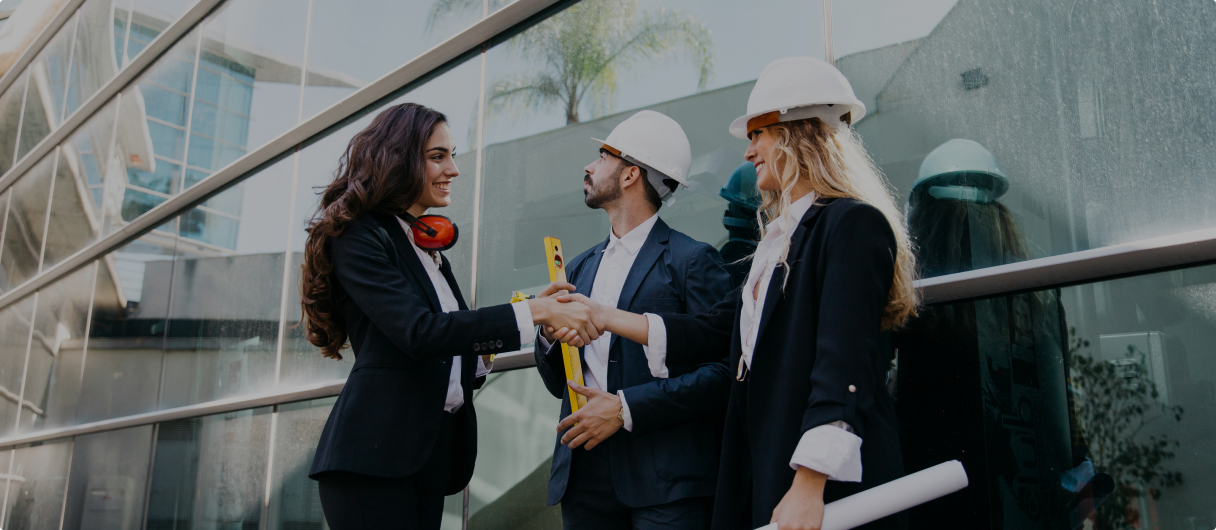 Three professionals in business attire and hard hats stand outside a modern glass building. Two of them, a man and a woman, are shaking hands, while the third woman holds blueprints. They appear to be engaged in a positive discussion, with a tape measure and safety gear visible, indicating a construction or engineering context.